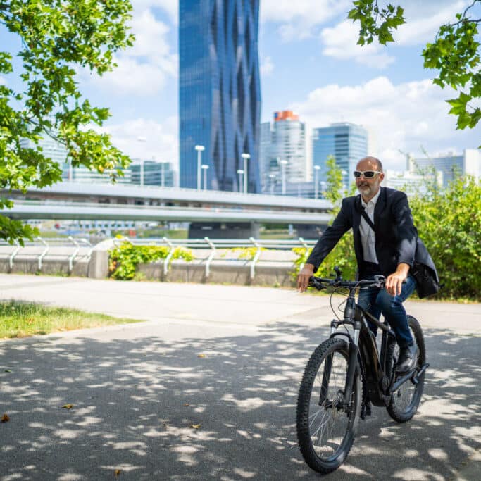friendly business man on e bike cycling through urban park on sunny summer day city buildings blurred in background